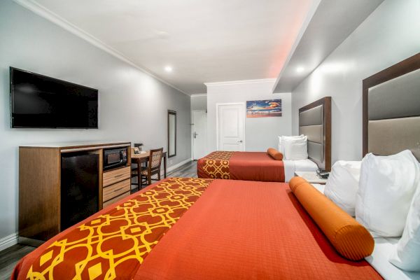 The image shows a hotel room with two beds, a flat-screen TV, a dresser, and a small table with chairs.