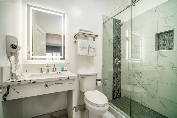 The image shows a modern bathroom with a shower, toilet, and a vanity. There's a backlit mirror and towels on a rack, creating a sleek look.