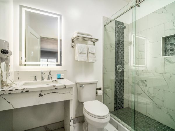 The image shows a modern bathroom with a shower, toilet, and a vanity. There's a backlit mirror and towels on a rack, creating a sleek look.
