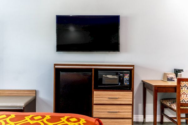 The image shows a hotel room setup with a wall-mounted TV, a microwave, a fridge, a small desk, and part of a bed with patterned bedding.