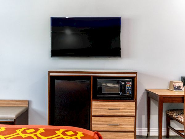 The image shows a hotel room setup with a wall-mounted TV, a microwave, a fridge, a small desk, and part of a bed with patterned bedding.