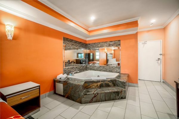 This image shows a room with an orange wall, a large mirror, a corner jacuzzi tub, a small dresser, and tiled flooring.