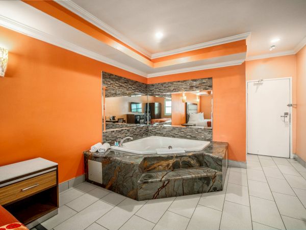 This image shows a room with an orange wall, a large mirror, a corner jacuzzi tub, a small dresser, and tiled flooring.