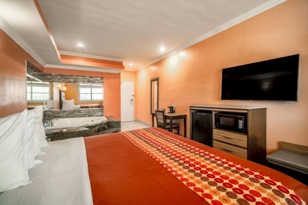 A hotel room with a bed, TV, microwave, and a hot tub near a large mirror. The walls are orange, and there's a polka dot bed runner.