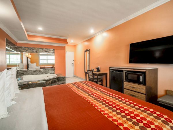 A hotel room with a bed, TV, microwave, and a hot tub near a large mirror. The walls are orange, and there's a polka dot bed runner.