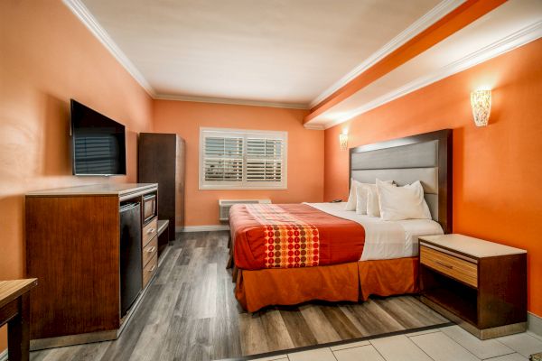 A hotel room with a double bed, TV, dresser, bedside tables, and orange walls. There's also a window with blinds and wood flooring.