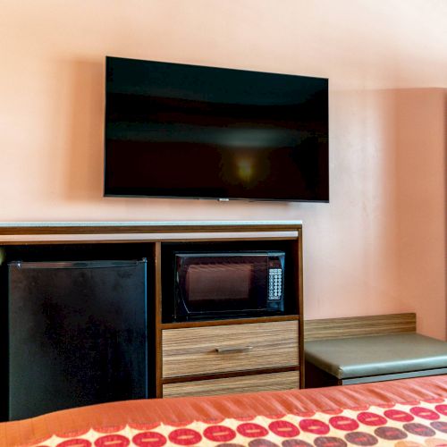 The image shows a hotel room interior with a wall-mounted TV, a small fridge, a microwave, and a partial view of a bed and wardrobe.