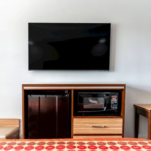 The image shows a hotel room with a wall-mounted TV, a cabinet with a mini-fridge and microwave, a table, and a chair.