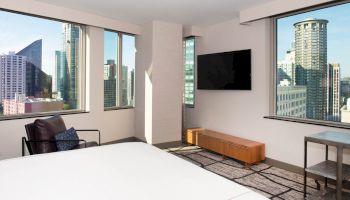 A modern hotel room with large windows showcasing a cityscape, a wall-mounted TV, and minimalistic furniture including a chair and table, is shown.
