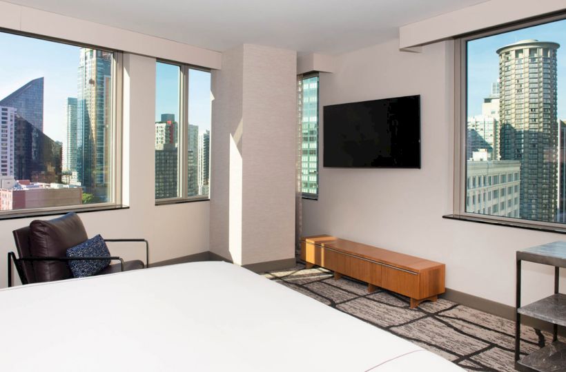 A modern hotel room with large windows showcasing a cityscape, a wall-mounted TV, and minimalistic furniture including a chair and table, is shown.