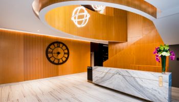 A modern lobby with a wooden spiral staircase, marble reception desk, wall clock, and flower arrangement.