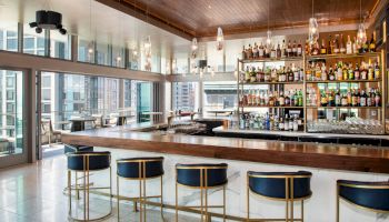 A modern bar with a variety of bottles on shelves, stylish barstools, pendant lights, and large windows overlooking a cityscape, ending the sentence.