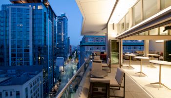 The image shows a modern balcony overlooking a cityscape at dusk, with seating and tables, adjacent to a well-lit indoor space.