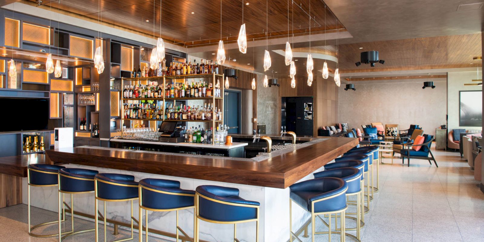 The image shows a modern bar with blue and gold stools, a well-stocked counter, hanging lights, and a comfortable seating area in the background.