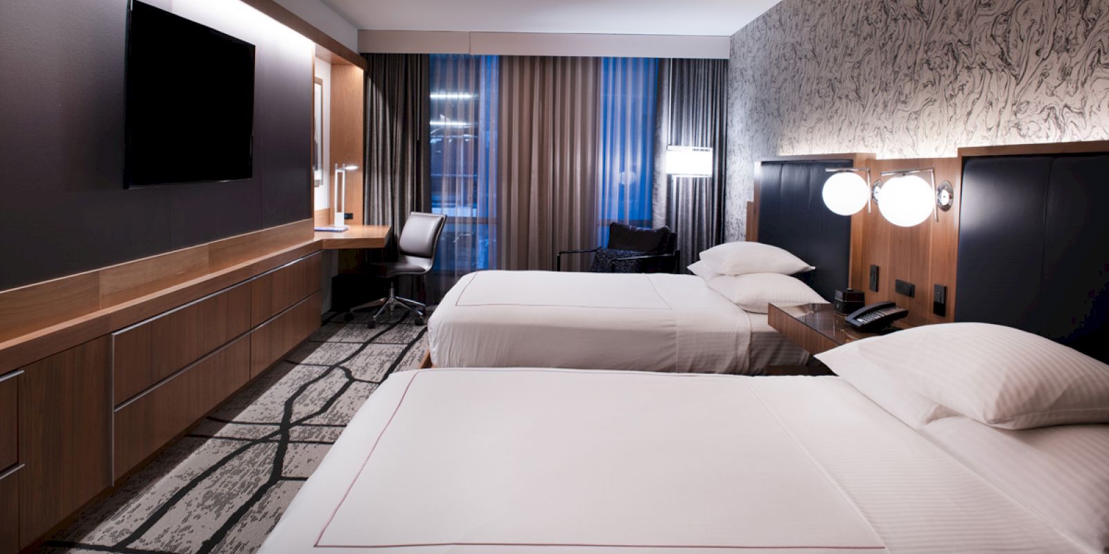 A modern hotel room with two beds, a flat-screen TV, a desk, and stylish lighting. The room has contemporary decor and a large window with blinds.