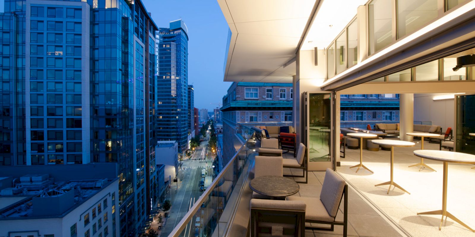 The image shows a modern cityscape at dusk, with tall buildings and a stylish balcony featuring chairs, tables, and outdoor lighting.