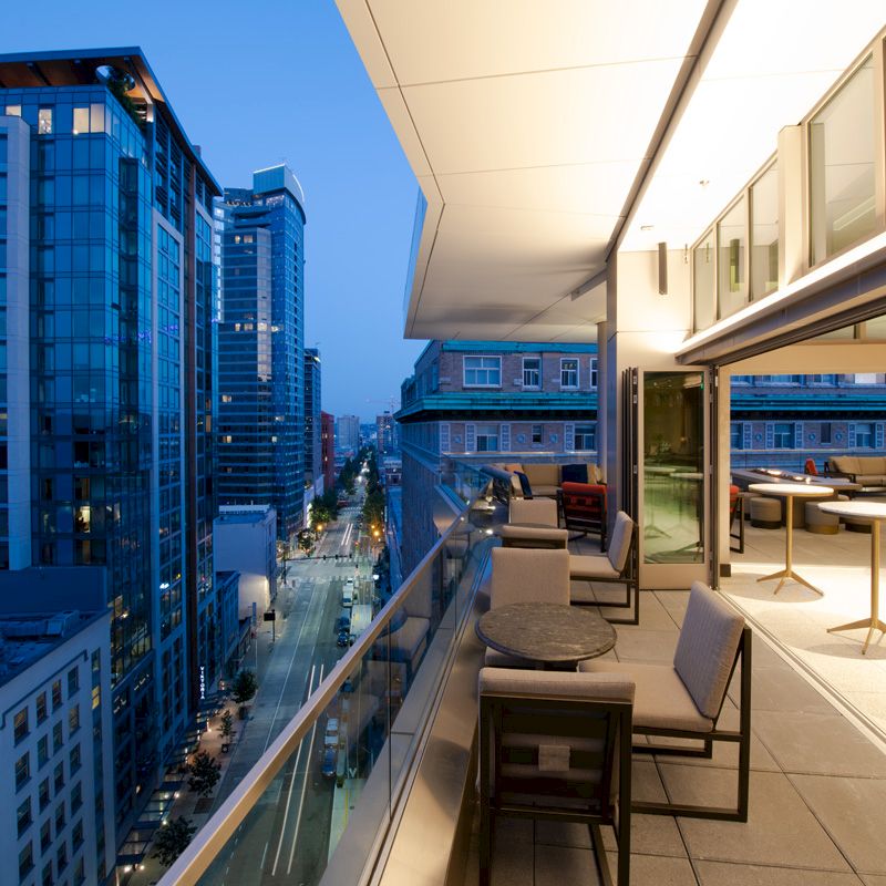 A modern rooftop balcony overlooks a city street with tall buildings. Outdoor seating and tables are arranged next to a glass barrier.