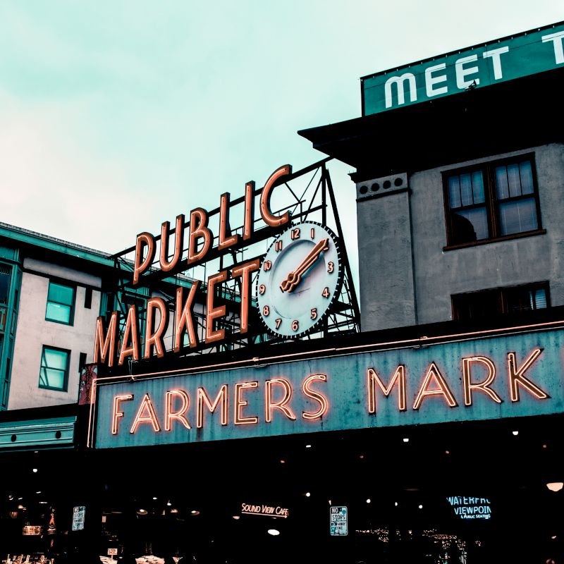 This image shows a public market with a neon sign that reads "Public Market" and "Farmers Market." A clock and other buildings are also visible in the background.