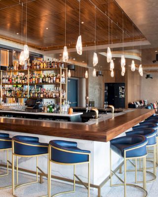 A modern bar with high-top stools, a variety of liquor bottles on shelves, hanging lights, and a lounge area in the background.