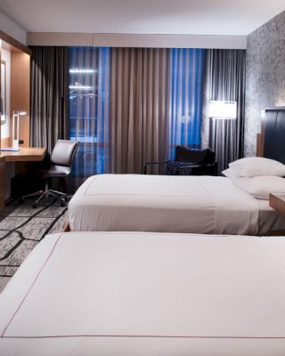 A modern hotel room features two neatly made beds, a wall-mounted TV, a work desk with a chair, and contemporary decor with stylish lighting.