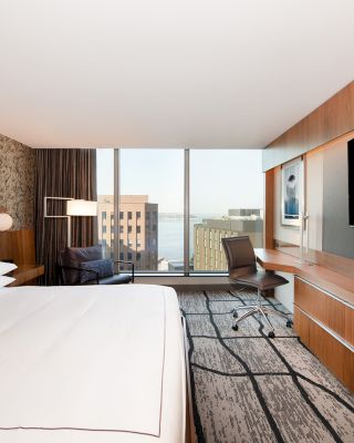 This image shows a modern hotel room with a large bed, a desk and chair, a flat-screen TV, and a window with a cityscape view.