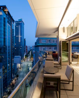 A modern urban balcony at twilight with outdoor seating, glass railings, and a panoramic city view with illuminated buildings.