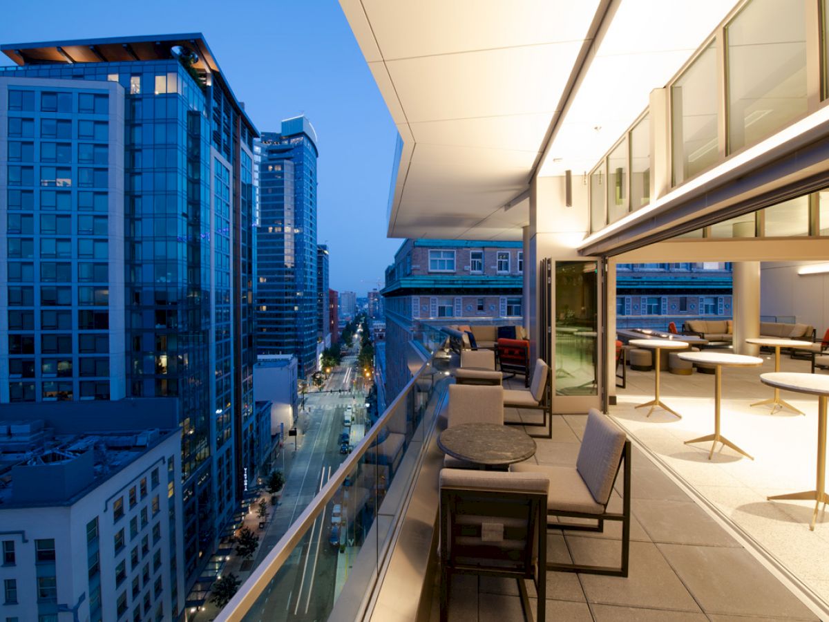 An urban rooftop balcony at dusk with modern seating, overlooking a bustling city street and tall buildings.