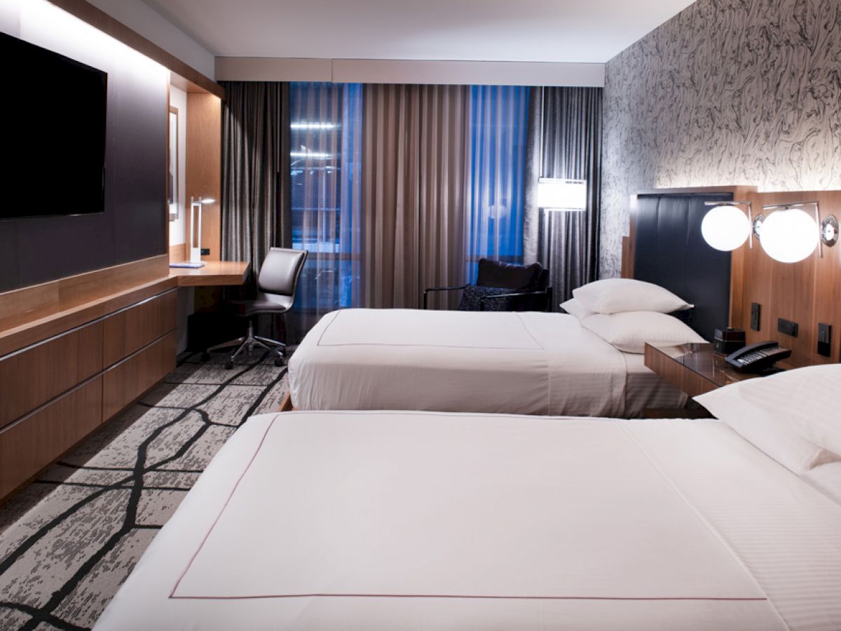 The image shows a modern hotel room with two neatly made beds, a wall-mounted TV, a desk, and seating area near the window.