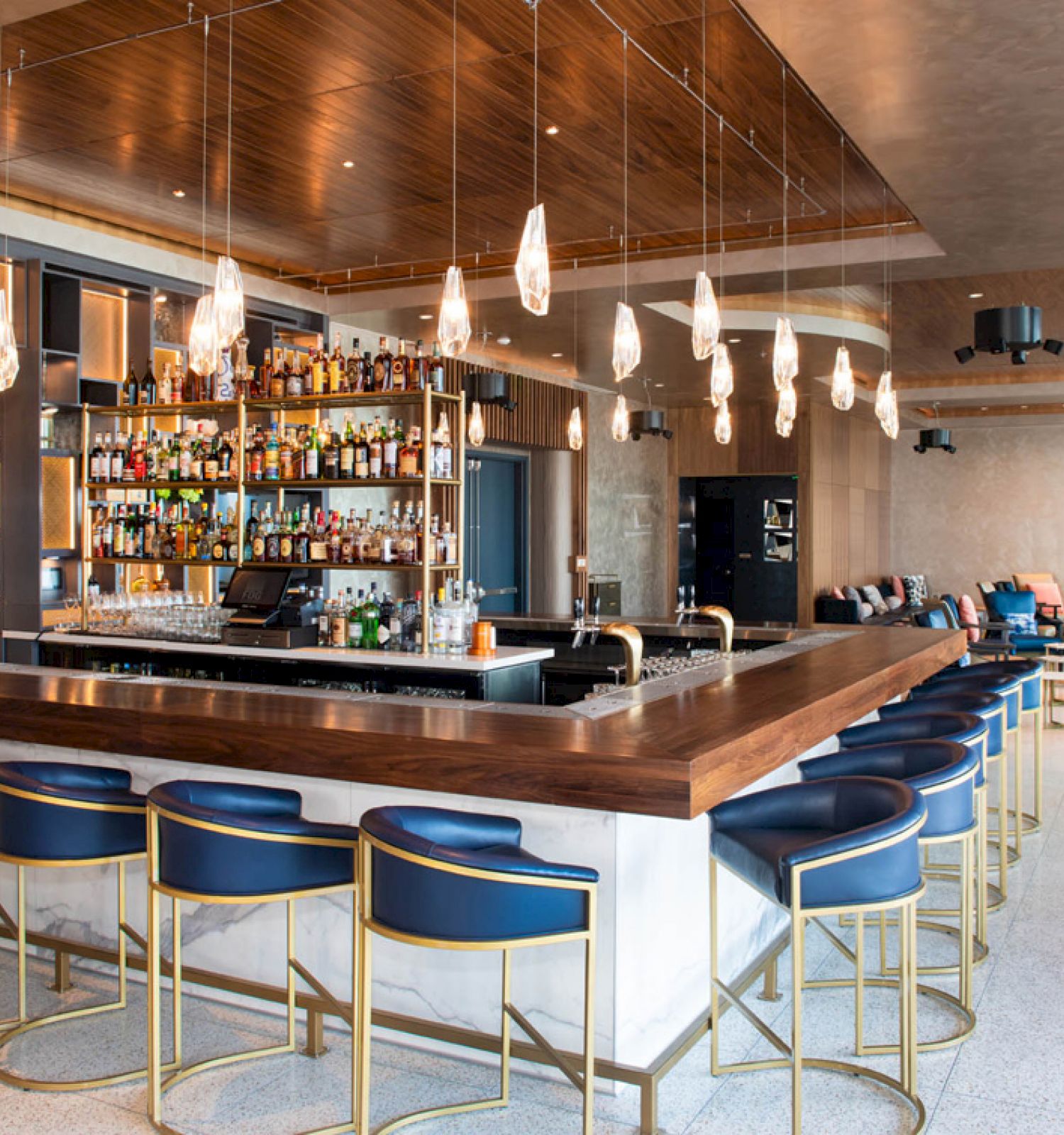The image shows a modern bar with blue bar stools, a wooden countertop, and a wide selection of bottles behind the counter.