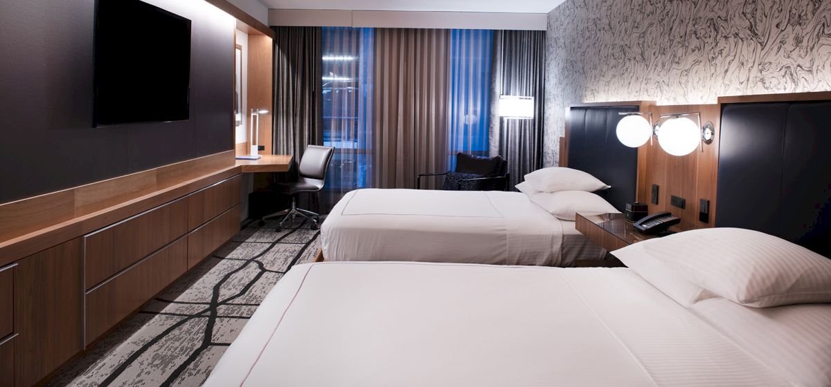 A modern hotel room features two neatly made beds, a flat-screen TV, a desk with a chair, and contemporary decor with accent lighting.