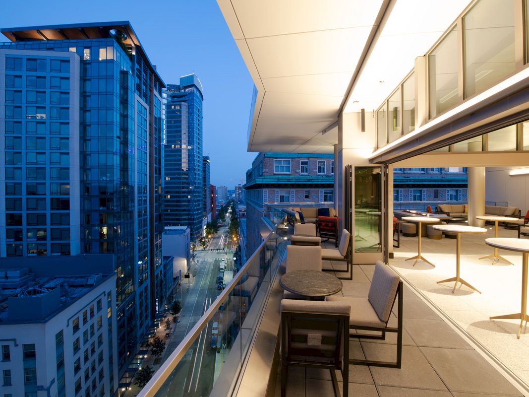 A rooftop terrace with seating and tables overlooks a street and tall buildings at dusk, with a clear view of the city skyline.