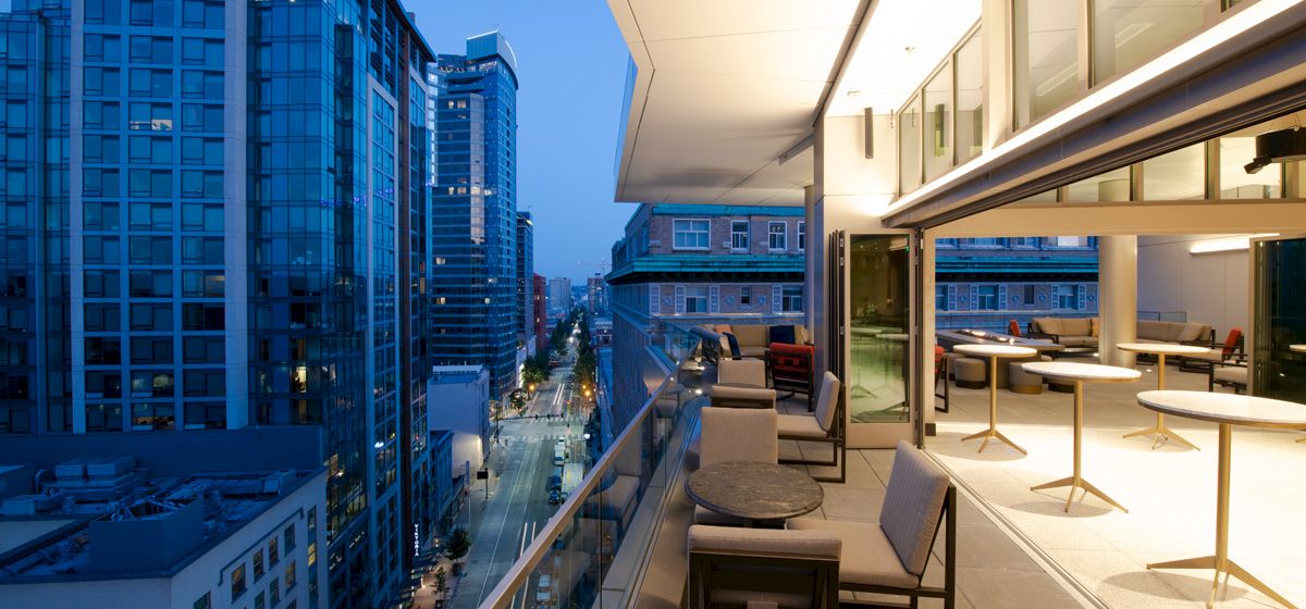 A rooftop terrace with seating and tables overlooks a street and tall buildings at dusk, with a clear view of the city skyline.