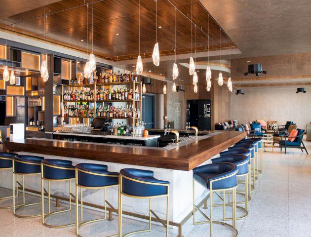 The image shows a modern bar with a large counter, blue bar stools, hanging lights, and a fully stocked back bar in a spacious and stylish setting.