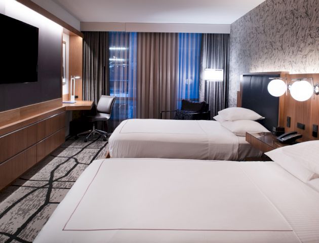 The image shows a modern hotel room with two double beds, a wall-mounted TV, a desk, and contemporary decor, featuring a geometric-patterned carpet.