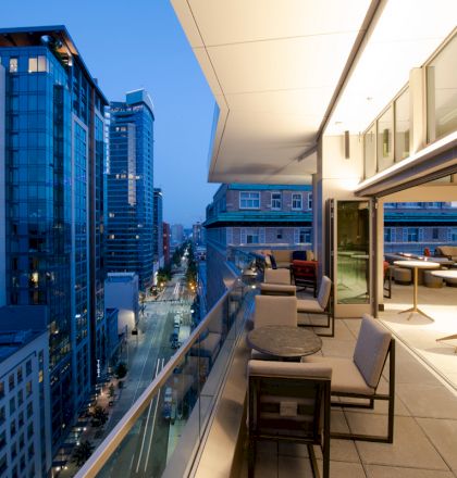 A modern balcony with seating overlooking a cityscape with tall buildings at dusk. The interior has tables and chairs with bright lighting.