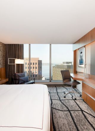 A modern hotel room features a large bed, a desk with chair, a wall-mounted TV, and a window with a city view and some buildings visible.
