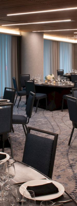 The image shows a neatly arranged banquet hall with round tables set with silverware, glassware, and black napkins. The room features elegant lighting.