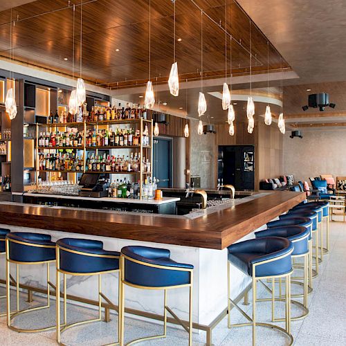 A modern bar with a wooden countertop, blue cushioned stools, and a well-stocked liquor shelf, featuring sleek lighting and a cozy seating area.