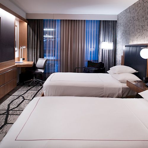The image shows a modern hotel room with two beds, a flat-screen TV, a desk with a chair, and contemporary decor and lighting fixtures on the wall.