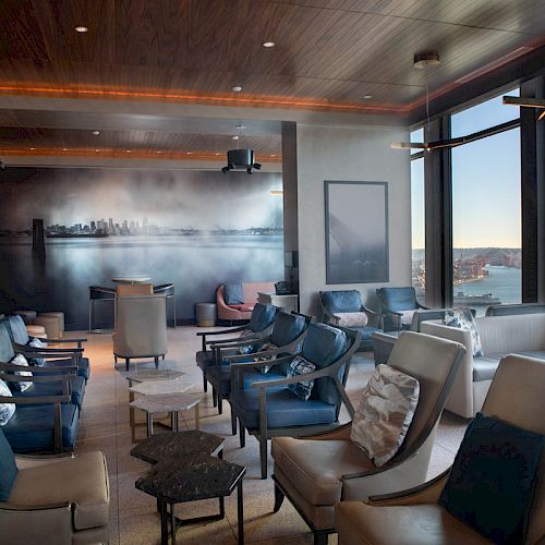 The image shows a modern lounge area with plenty of seating, a large mural, and floor-to-ceiling windows offering a view of the waterfront.