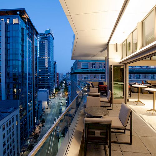 A modern cityscape with tall buildings is shown at twilight, alongside an elegant balcony with tables and chairs for outdoor seating.