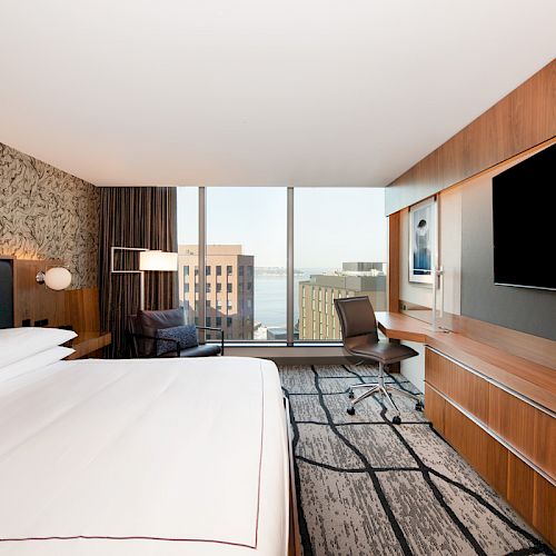 A modern hotel room with a large bed, wall-mounted TV, desk, chair, and a window offering a city view with buildings in the background.