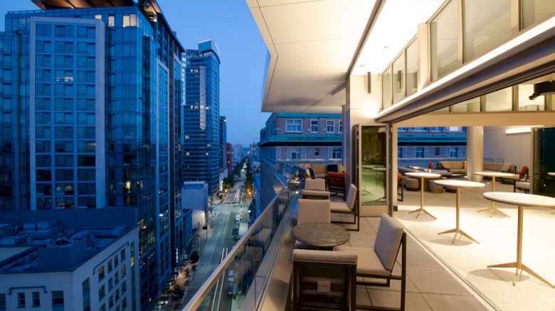 The image shows a modern balcony overlooking a cityscape with tall buildings, featuring outdoor seating and tables, illuminated in the evening.