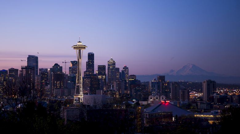 The image shows the Seattle skyline at dusk, featuring the Space Needle prominently, with city buildings and Mount Rainier in the background.