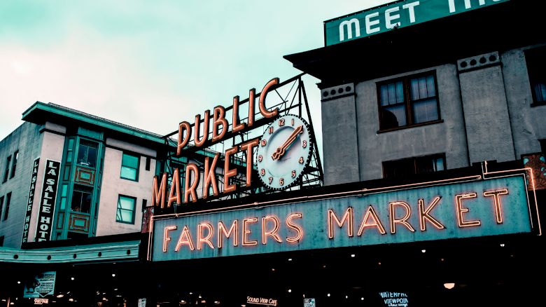 The image shows an iconic public market with neon signs for a "Farmers Market," a clock, and surrounding buildings with vintage architecture.