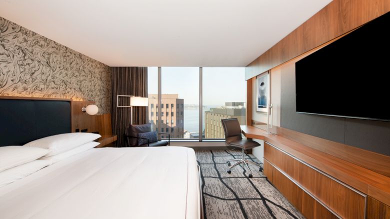 A modern hotel room with a king-sized bed, desk, large TV, and a city view through floor-to-ceiling windows, featuring stylish decor and furnishings.