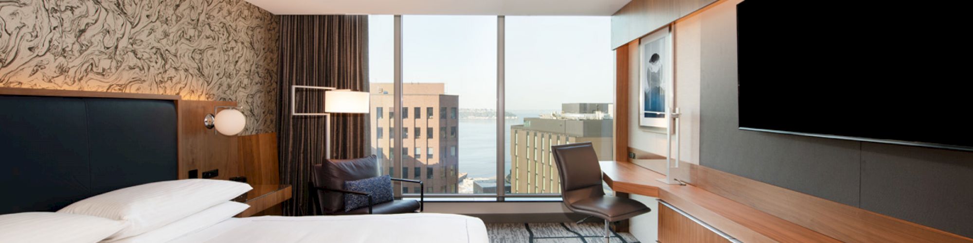 A modern hotel room with a large bed, wall-mounted TV, desk, armchair, and a window view of buildings and water outside.