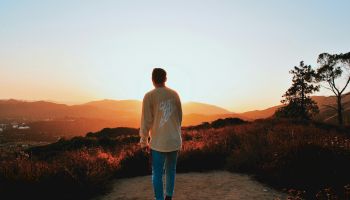 A person stands on a path, gazing at a mountain view during sunset, surrounded by trees and a glowing sky.