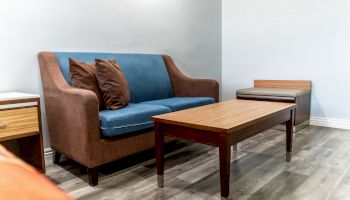 The image shows a room with a brown and blue sofa, two brown cushions, a wooden coffee table, an end table, and a wooden floor.