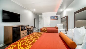 The image shows a hotel room with two double beds, a wall-mounted TV, a desk, and a dresser, all in a modern setting.